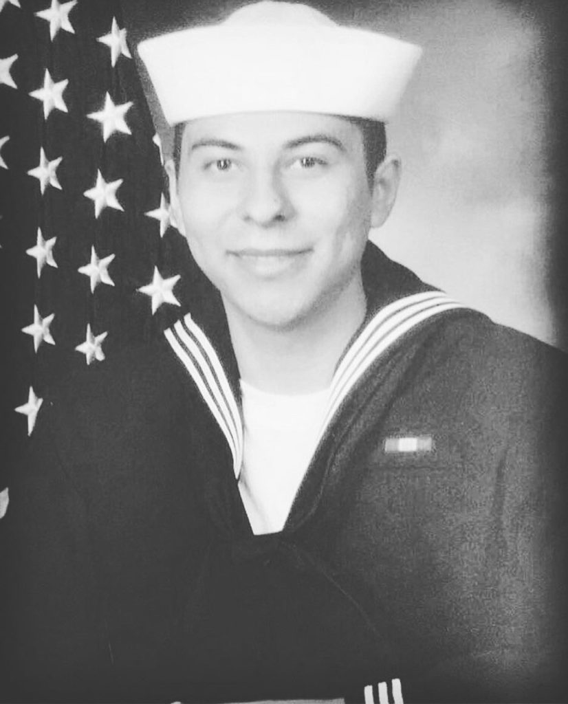 Portrait of a smiling U.S. Navy sailor in uniform, standing in front of an American flag.