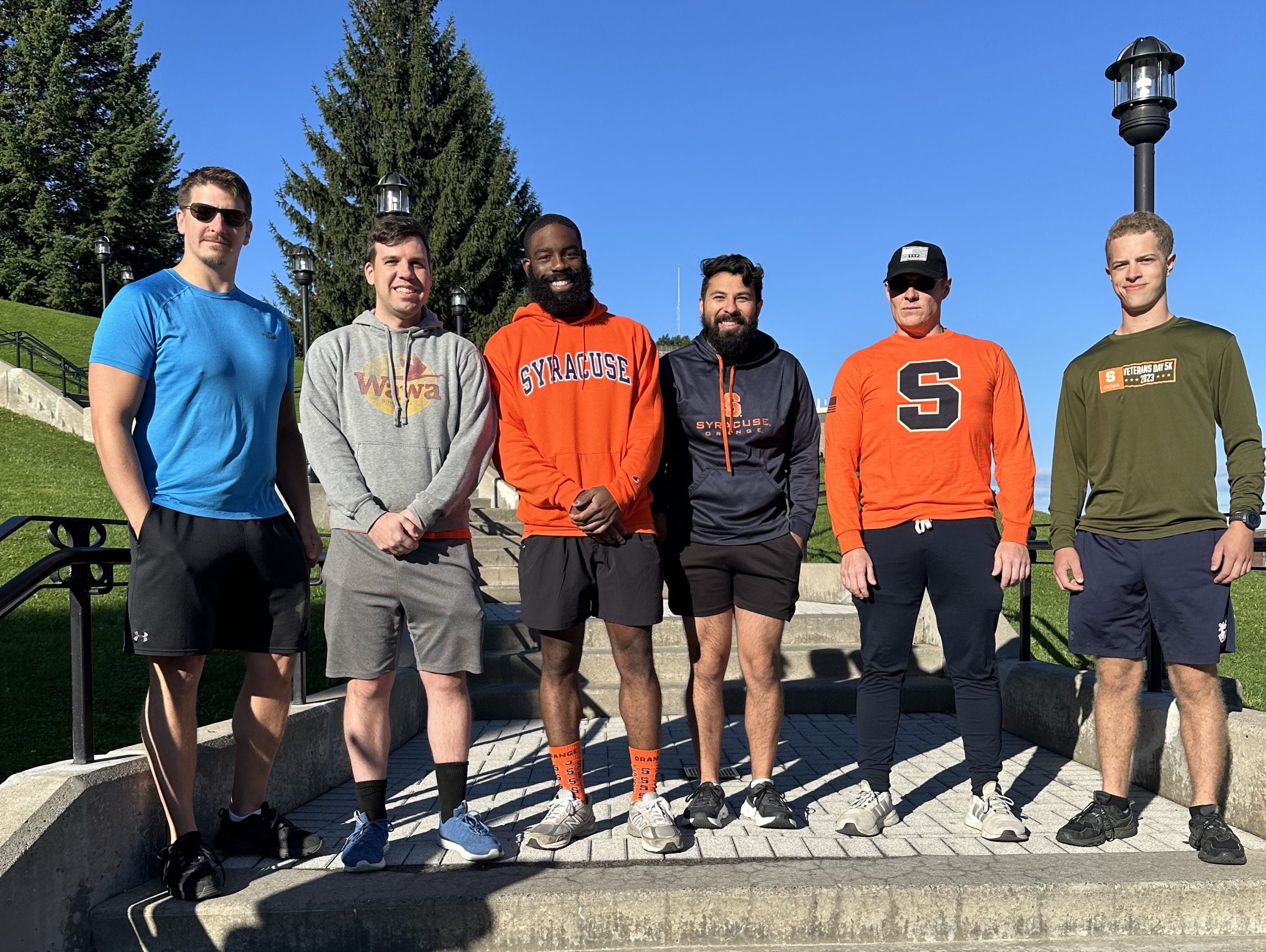 Six individuals standing on steps outdoors, wearing various casual and athletic clothing with collegiate logos such as "Syracuse." Some are smiling, and the setting includes green grass and a clear blue sky.