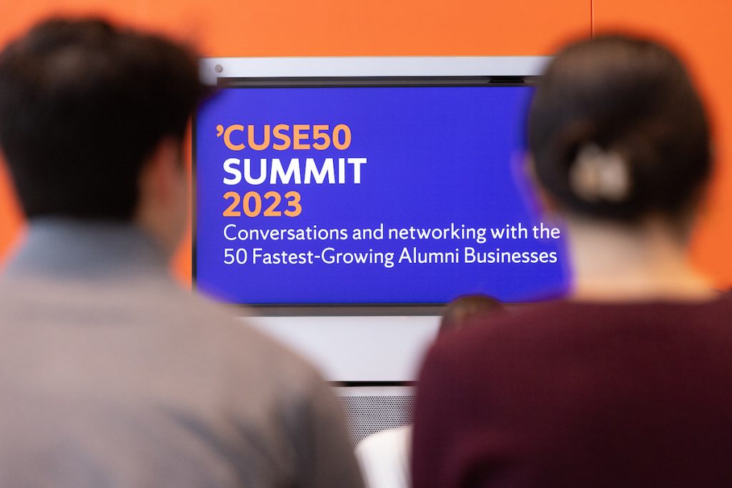 Two individuals seated, viewing a screen displaying 'CUSE50 Summit 2023' with a subtitle about conversations and networking with the 50 fastest-growing alumni businesses.