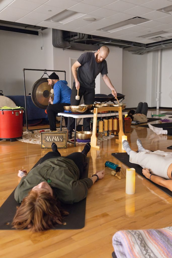 Two individuals participate in a sound therapy session with a therapist playing various percussion instruments including gongs and singing bowls.