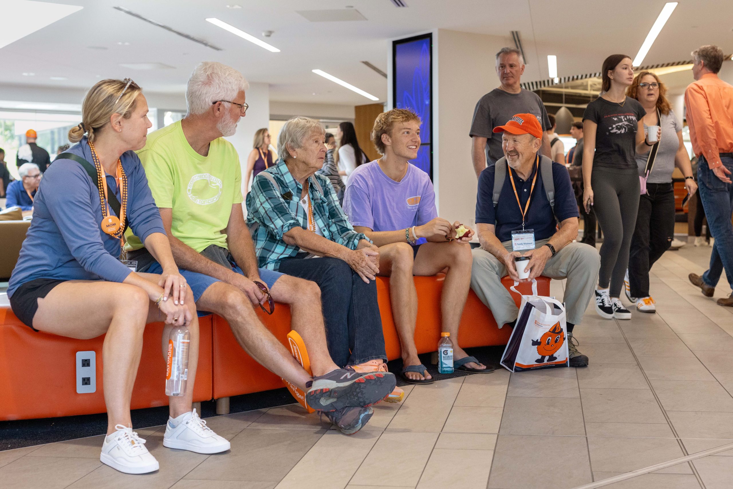 Five people sitting on an orange bench in a crowded lobby, wearing casual attire and accessories like lanyards and hats, engaged in conversation and laughter.