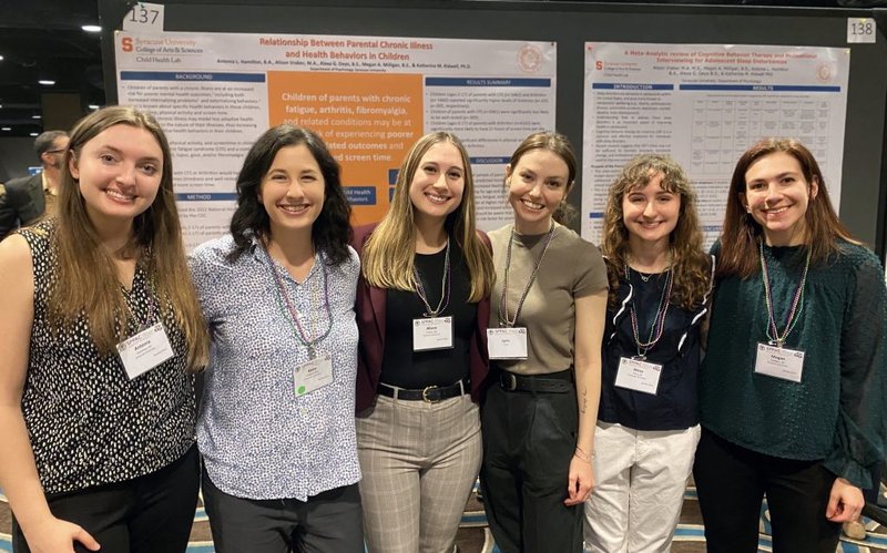 Six individuals smiling at a scientific conference, standing in front of posters that discuss health studies. Each person is wearing a badge.