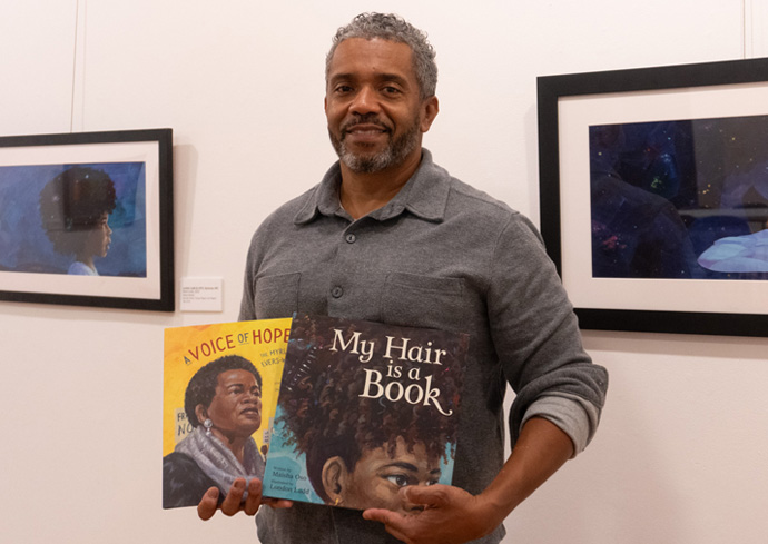 A man stands smiling in an art gallery, holding two picture books titled "A Voice of Hope" and "My Hair is a Book." Behind him are framed artworks on a white wall.