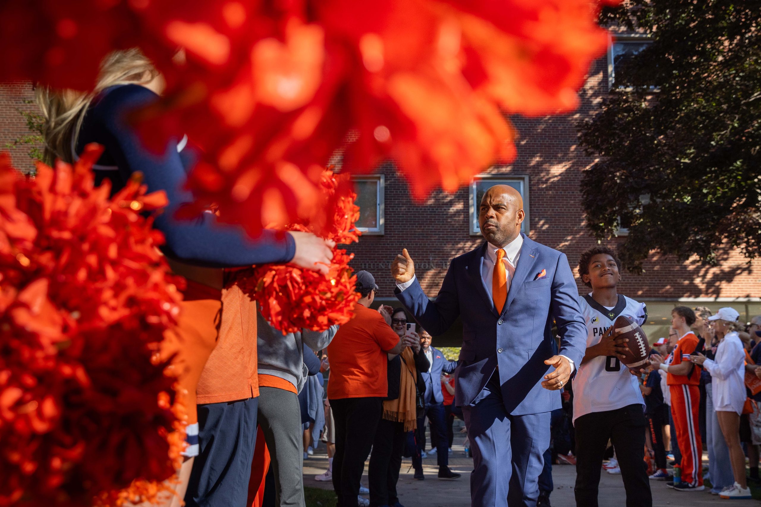 Person in a suit, waving while walking through a festive crowd with other people holding orange pompoms.