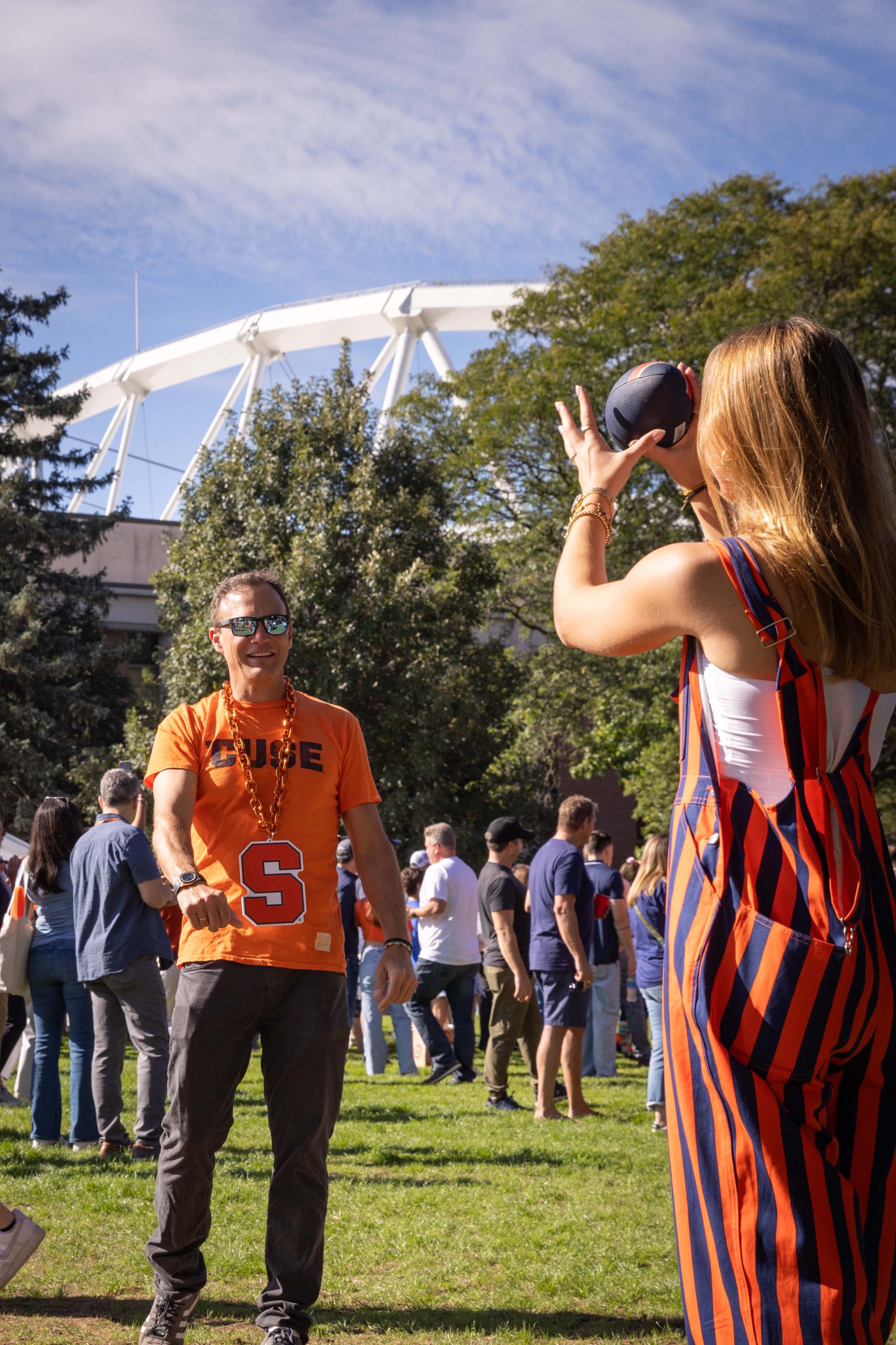 A person wearing a Syracuse University T-shirt playing catch with another individual in a striped outfit, with a group of people and trees in the background under a clear blue sky.