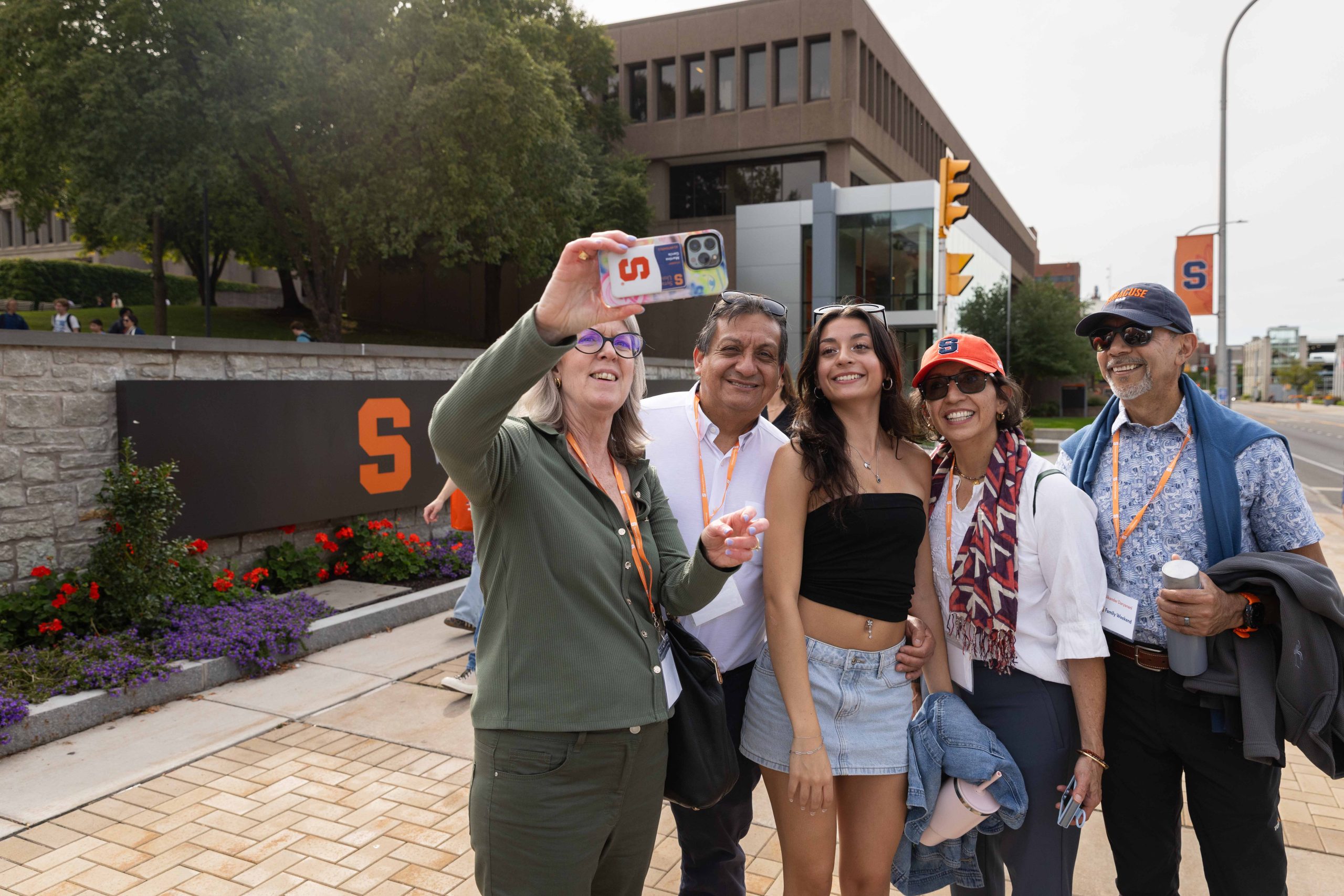 Group of five people taking a selfie in front of Syracuse University, with orange-themed clothing and accessories, expressing joy.