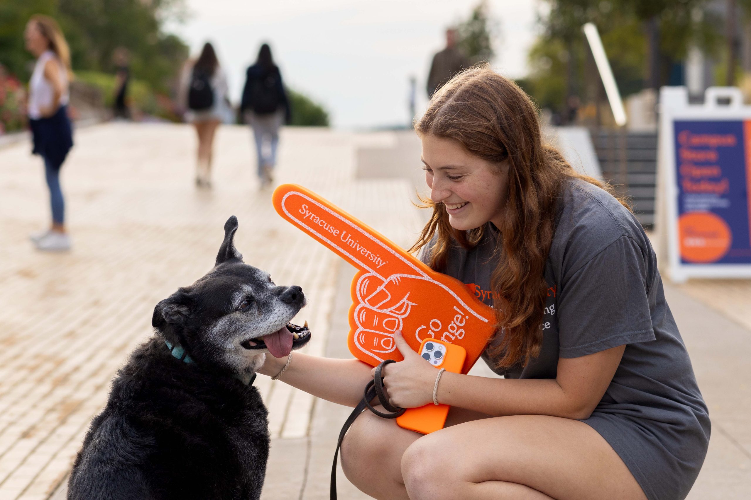 A person smiling at a black dog while holding a Syracuse University foam finger, outdoors on a campus walkway with other pedestrians in the background.