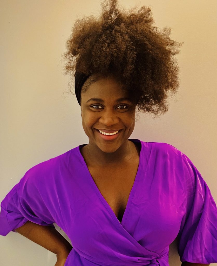 Person smiling in a bright purple top, with a voluminous hairstyle, against a light background.