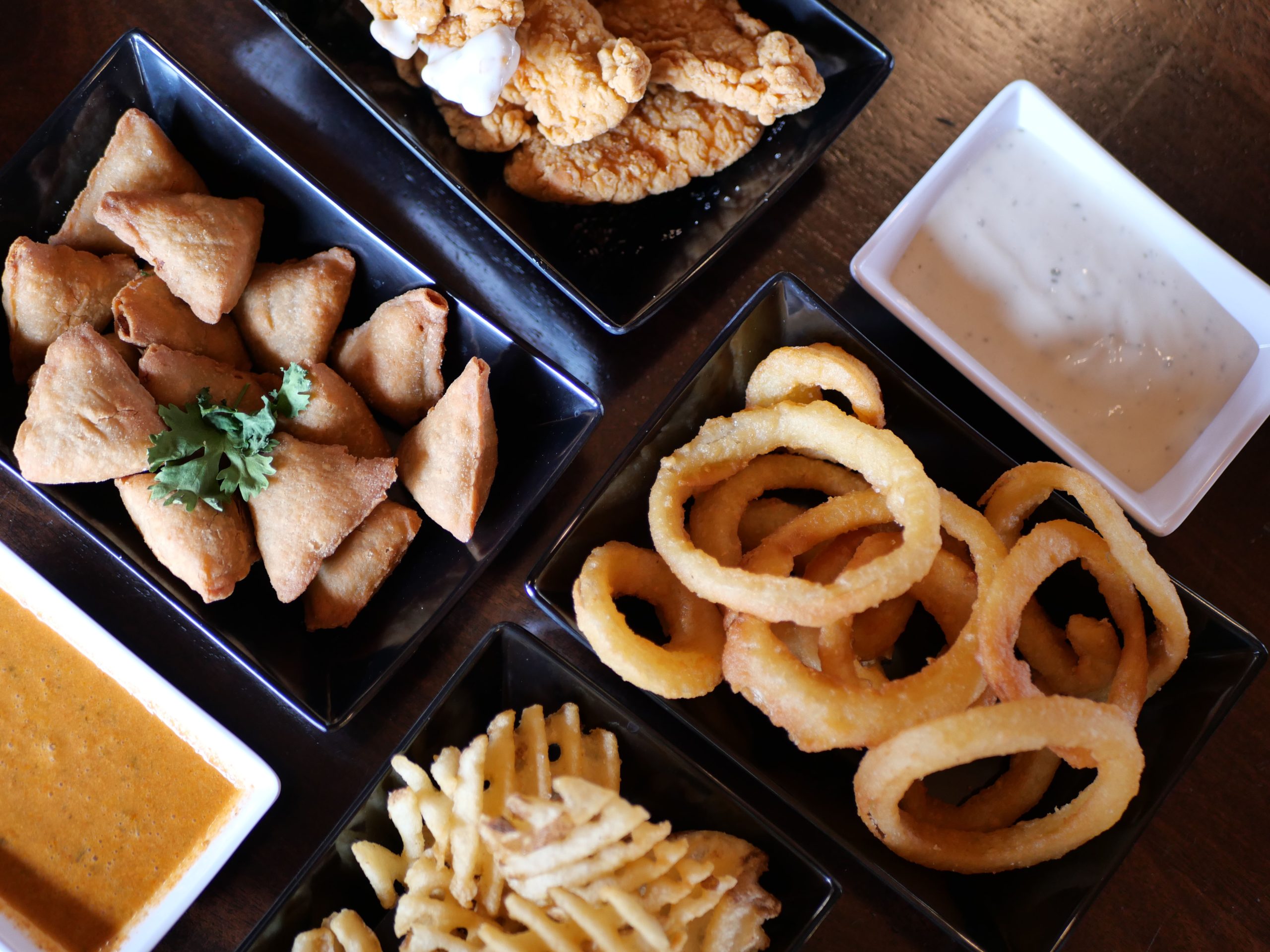 A variety of appetizers on a wooden table, including samosas, crispy chicken, onion rings and French fries served with dipping sauces.