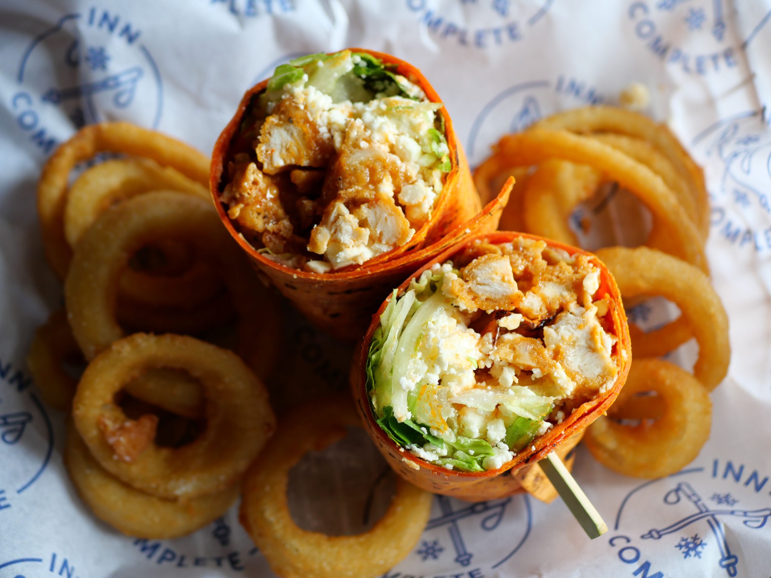 A halved wrap filled with chicken and vegetables, served with a side of onion rings on a paper marked with the logo of Inn Complete.