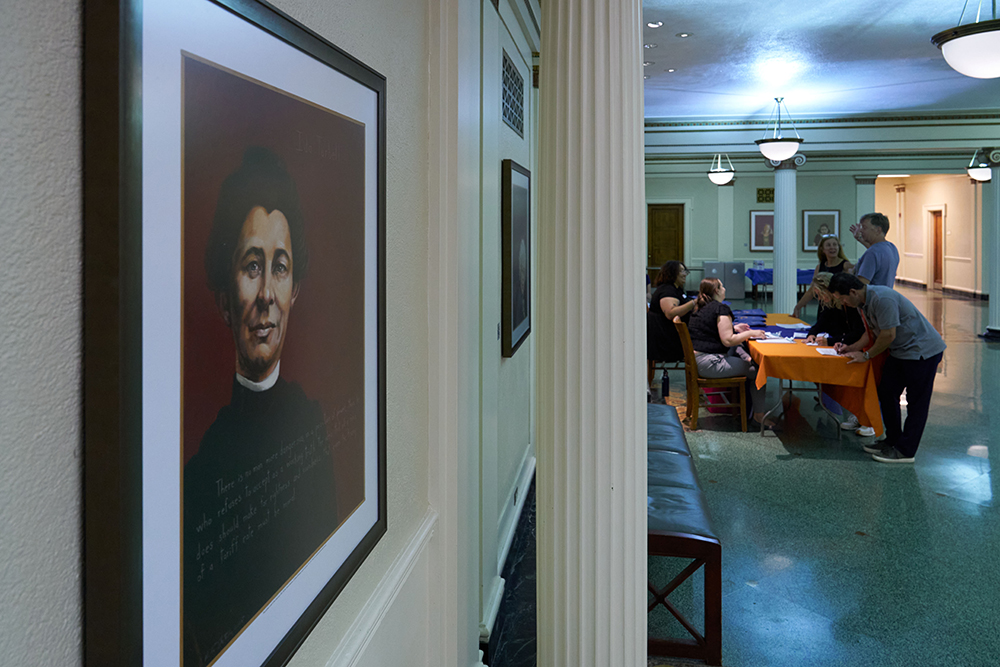 A framed portrait hangs on a wall in a hallway. In the background, several people gather around a table with an orange tablecloth, engaging in conversation. The area is well-lit with classic overhead lights.