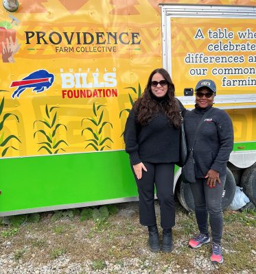 Two individuals standing in front of a colorful Providence Farm Collective trailer. The trailer features a Buffalo Bills Foundation logo and corn graphics. Both people are smiling and dressed in casual clothing.