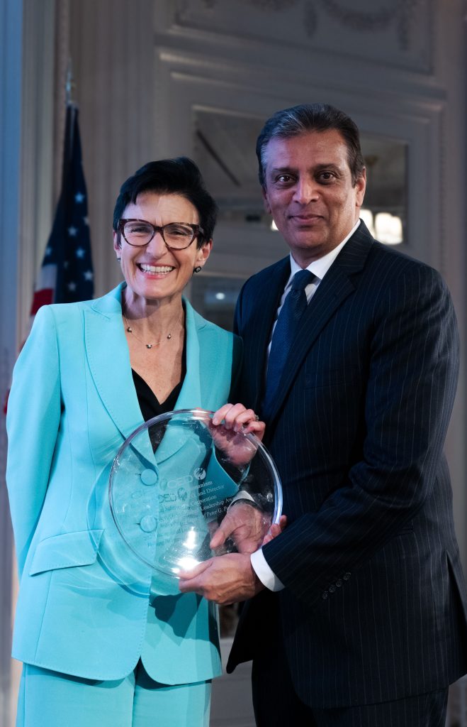 Two individuals holding an award at an event, with the U.S. flag in the background. The person on the left is wearing a light blue suit and glasses, while the person on the right is dressed in a dark suit.