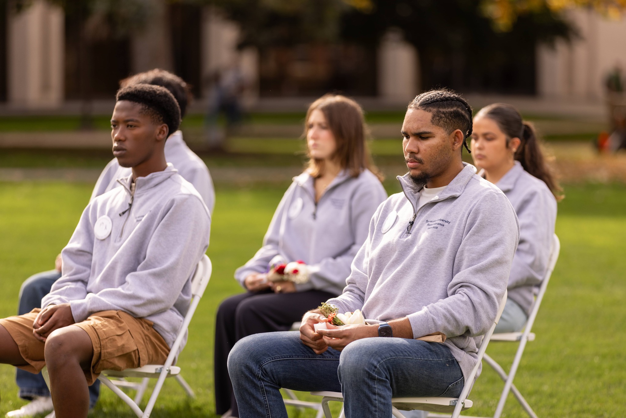 Four individuals seated outdoors in a row on white chairs, each wearing gray sweatshirts.