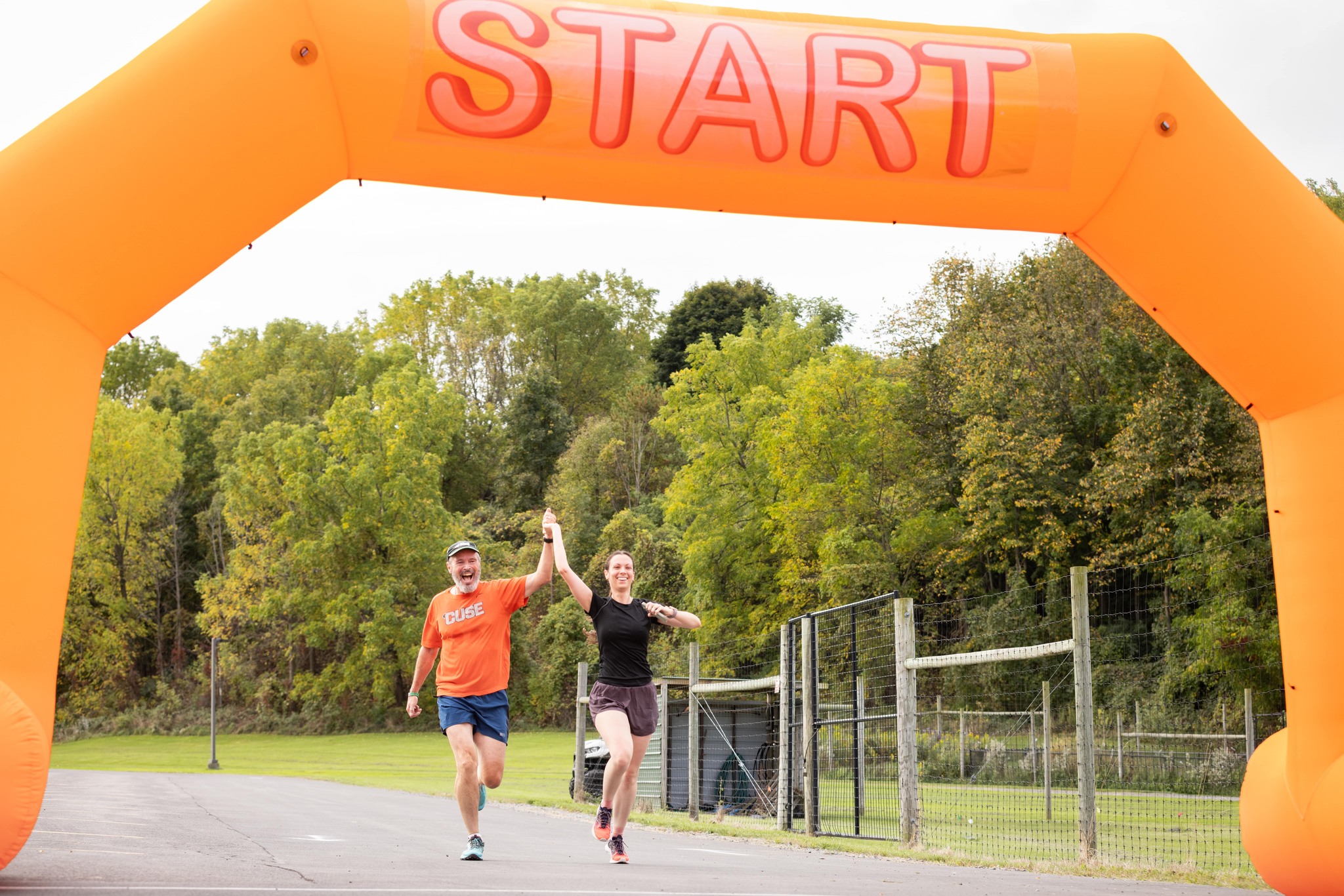 Two runners crossing under a large orange inflatable arch labeled "START" in a park setting.