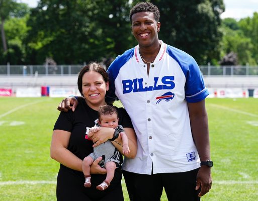 A smiling family stands on a football field. A woman holds a baby, and a tall man wearing a "BILLS" shirt has an arm around her. Trees and bleachers are visible in the background.