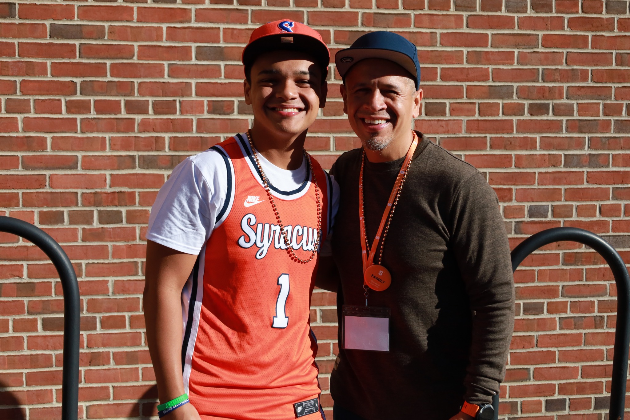 Two individuals smiling in front of a brick wall, one wearing a Syracuse basketball jersey and the other in a brown sweater and matching cap.