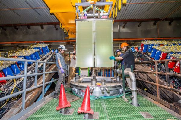 Two workers are installing a large, vertical metal panel into a complex machine setup. Cones and tools are visible around them on the floor, and numerous cables and mechanical components surround the area.