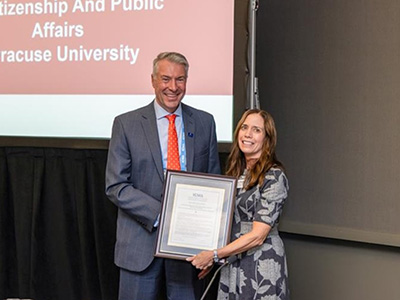 Two people are standing in a conference room in front of a digital screen. One is holding a framed certificate.