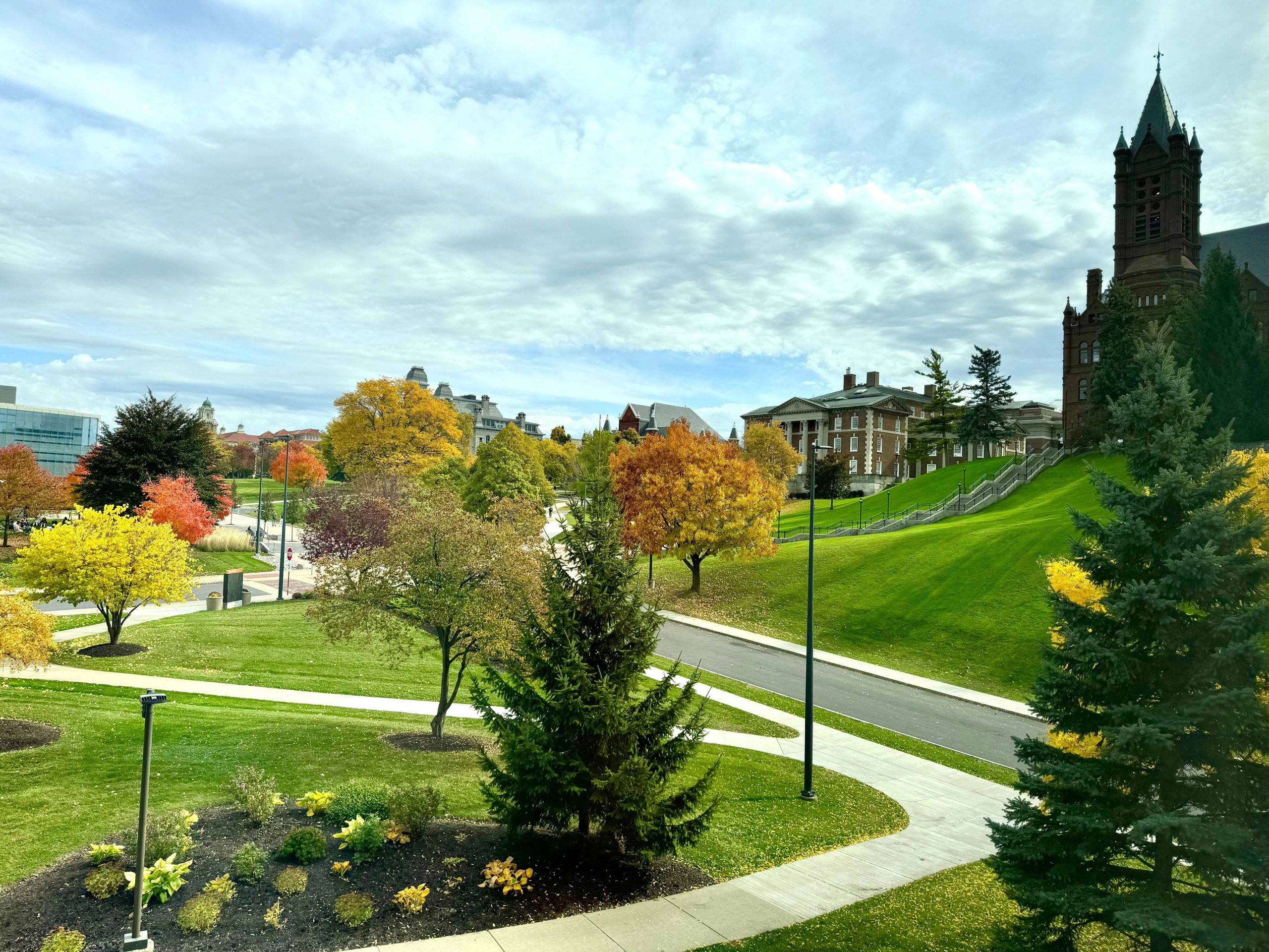 View of a landscaped campus with vibrant autumn foliage. A winding pathway leads towards a prominent historic building with a clock tower under a partly cloudy sky.