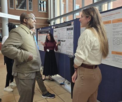 man in tan jacket speaks to a young woman presenting her research poster