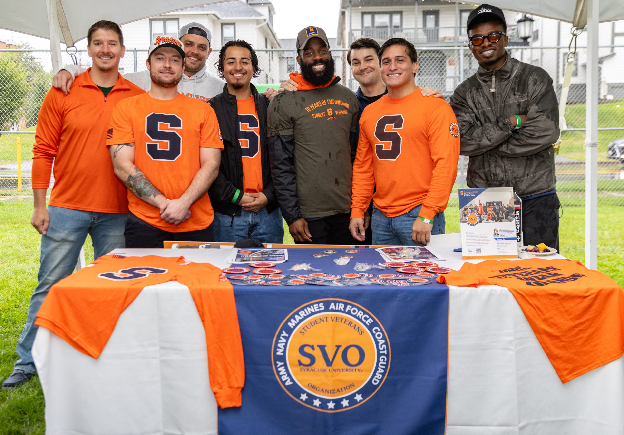 Group of eight people smiling behind a table with a banner that reads "Syracuse University Student Veterans Organization (SVO)" and "Student Veterans, America" at an outdoor event. They are wearing Syracuse University apparel.