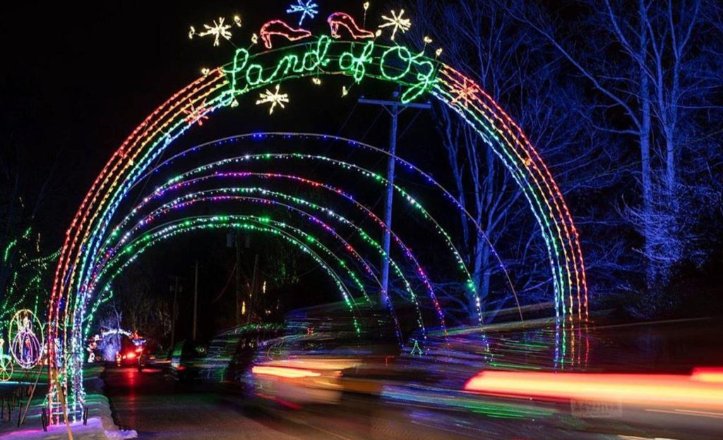 Night view of a colorful illuminated archway with the text 'Land of Oz' at a festive lights display, with motion-blurred vehicle lights passing underneath.