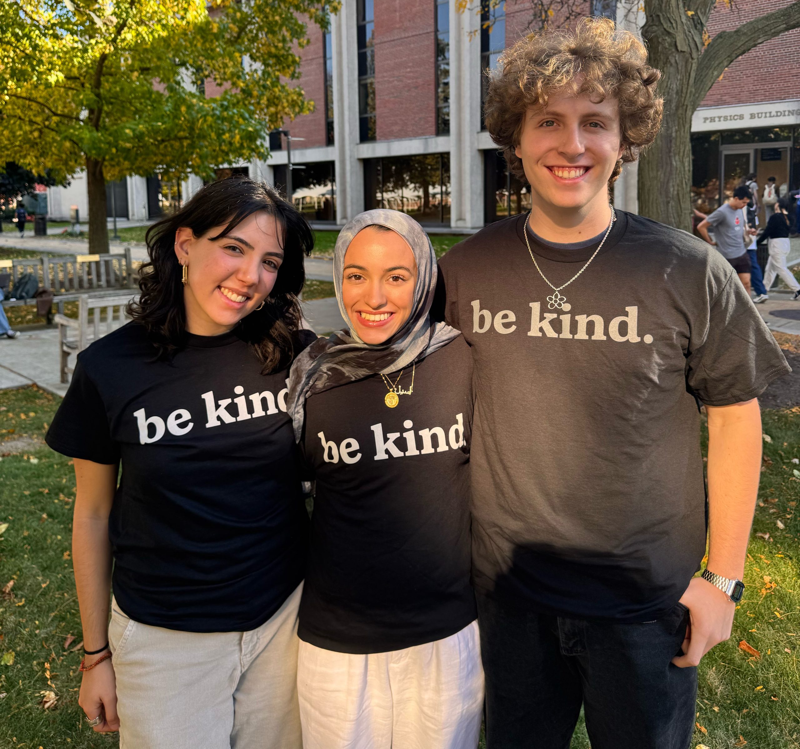 Three people wearing 'be kind.' t-shirts, standing together and smiling in a campus setting with trees and a building in the background.