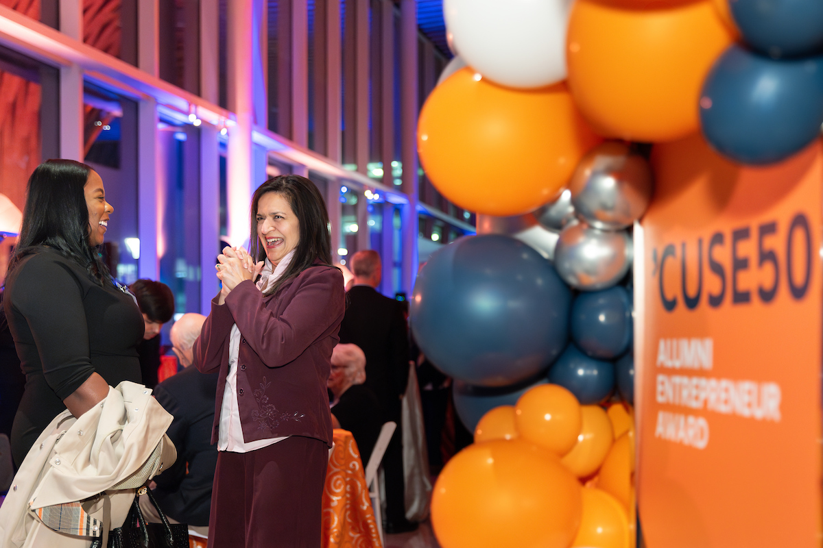 Two individuals are engaged in a lively conversation at the 'Cuse 50 Alumni Entrepreneur Award event, surrounded by colorful balloons and vibrant lighting.