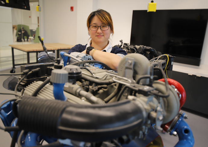 A professor poses with car parts inside of a classroom.