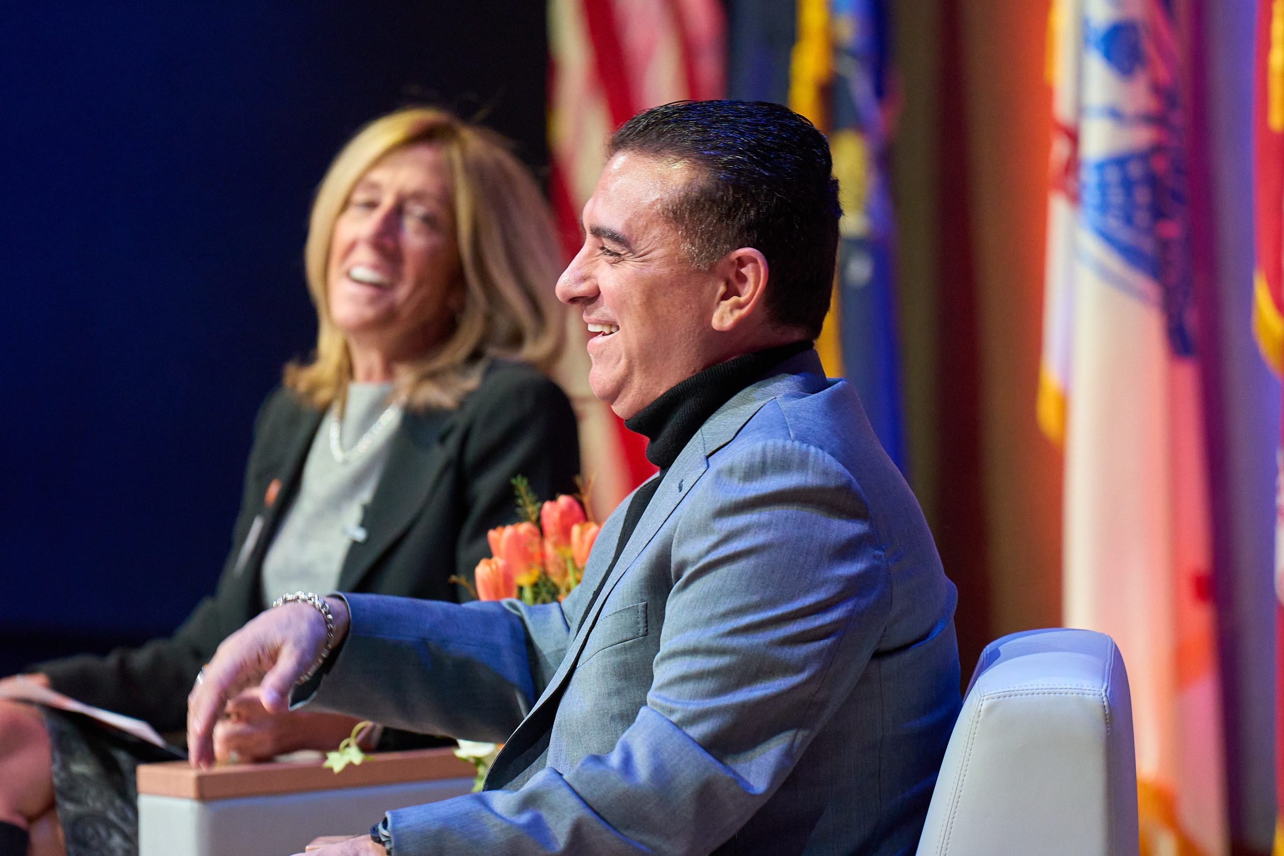 Two individuals are smiling while sitting at a panel discussion during an event. The person on the right is wearing a gray suit.