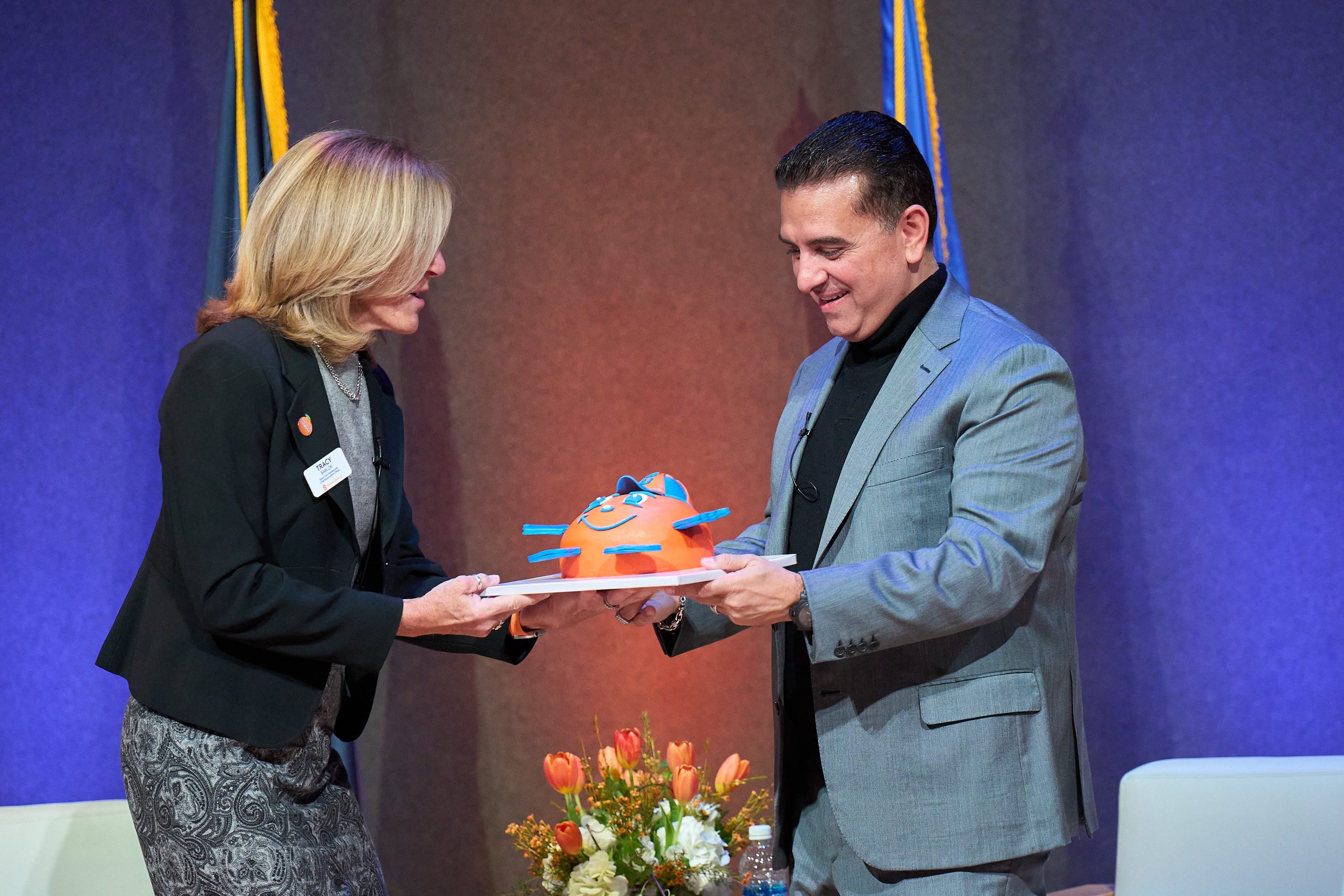 Two individuals exchanging an orange cake at a formal event.