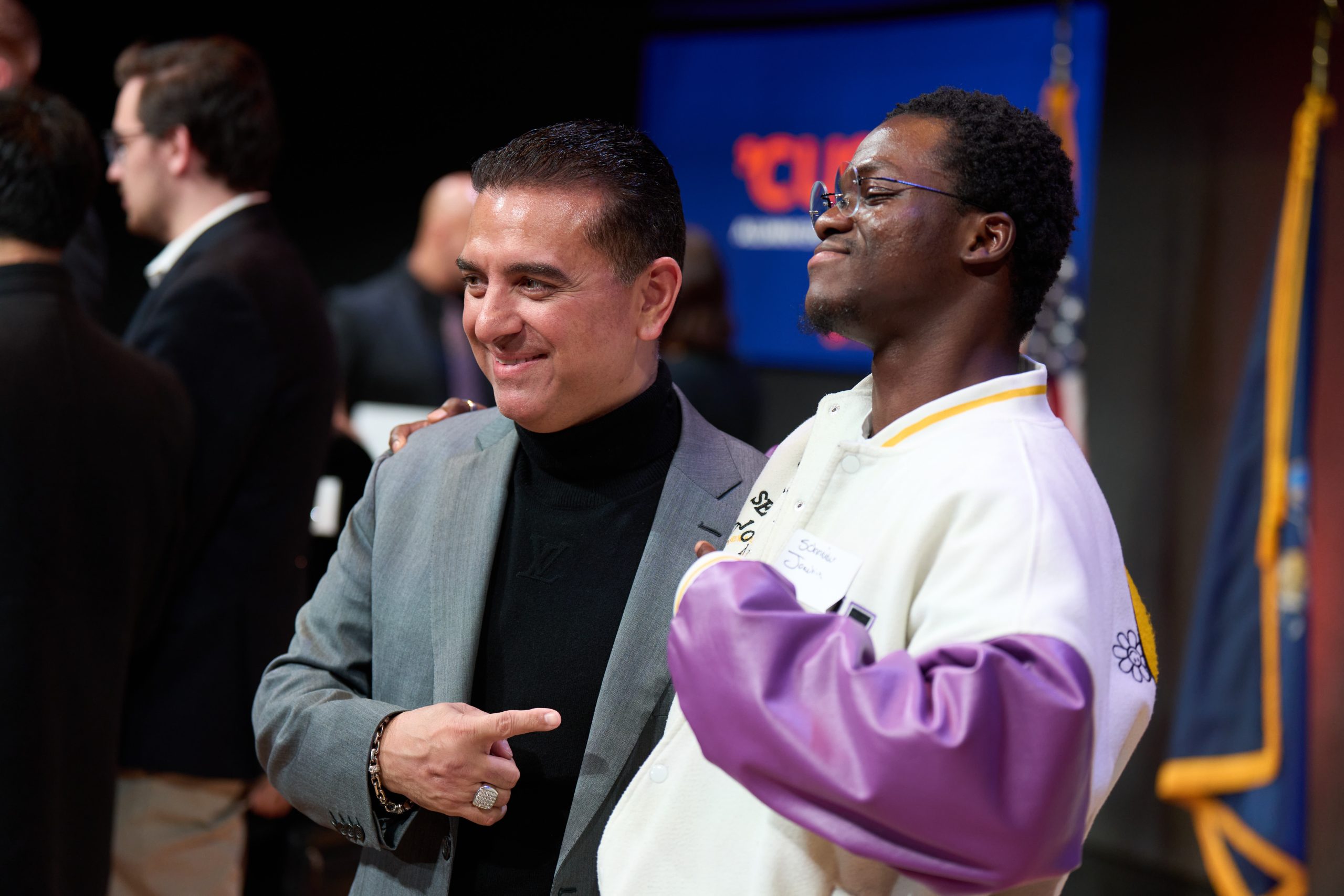 Two individuals posing together while smiling at an event. One is wearing a gray blazer and the other is dressed in a purple and white sports jacket.