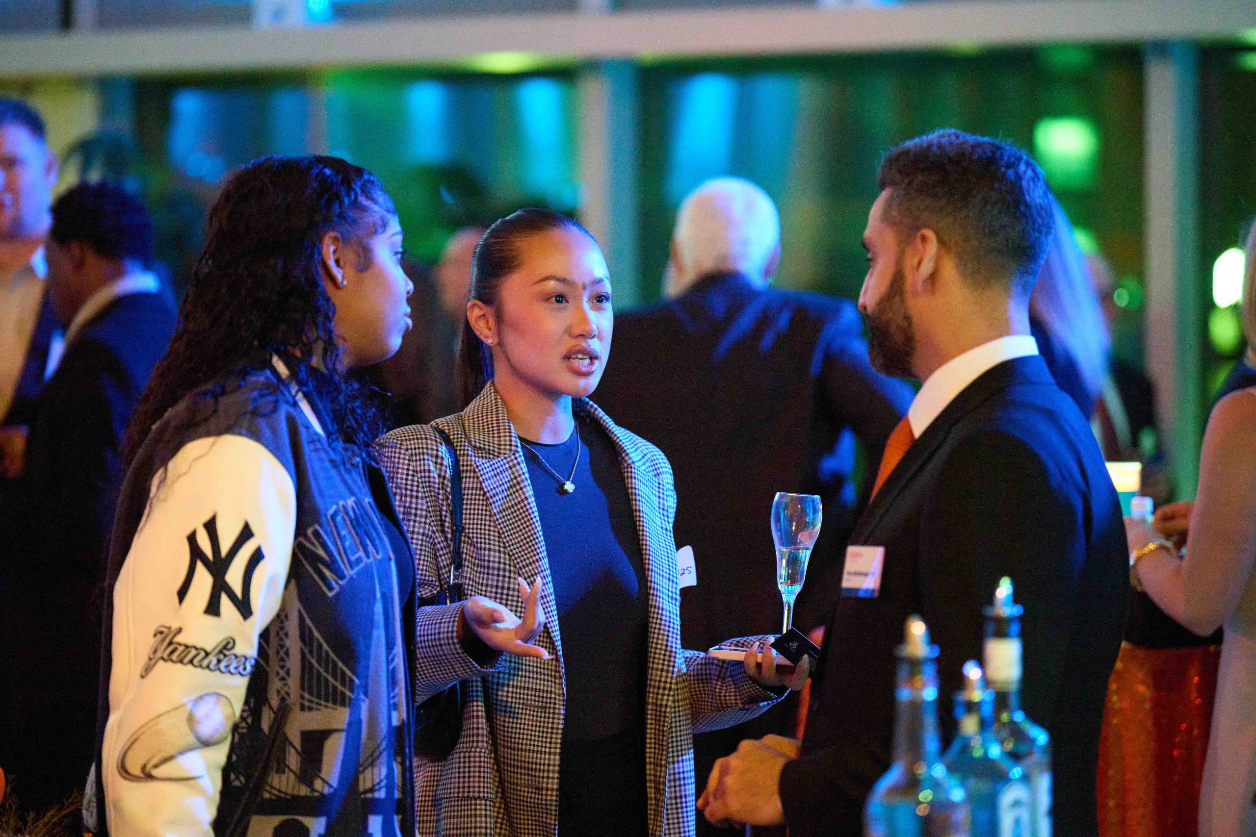 Three individuals engaging in conversation at a networking event, with one person holding a glass of wine. The environment is vibrant with colorful lighting and other attendees in the background.