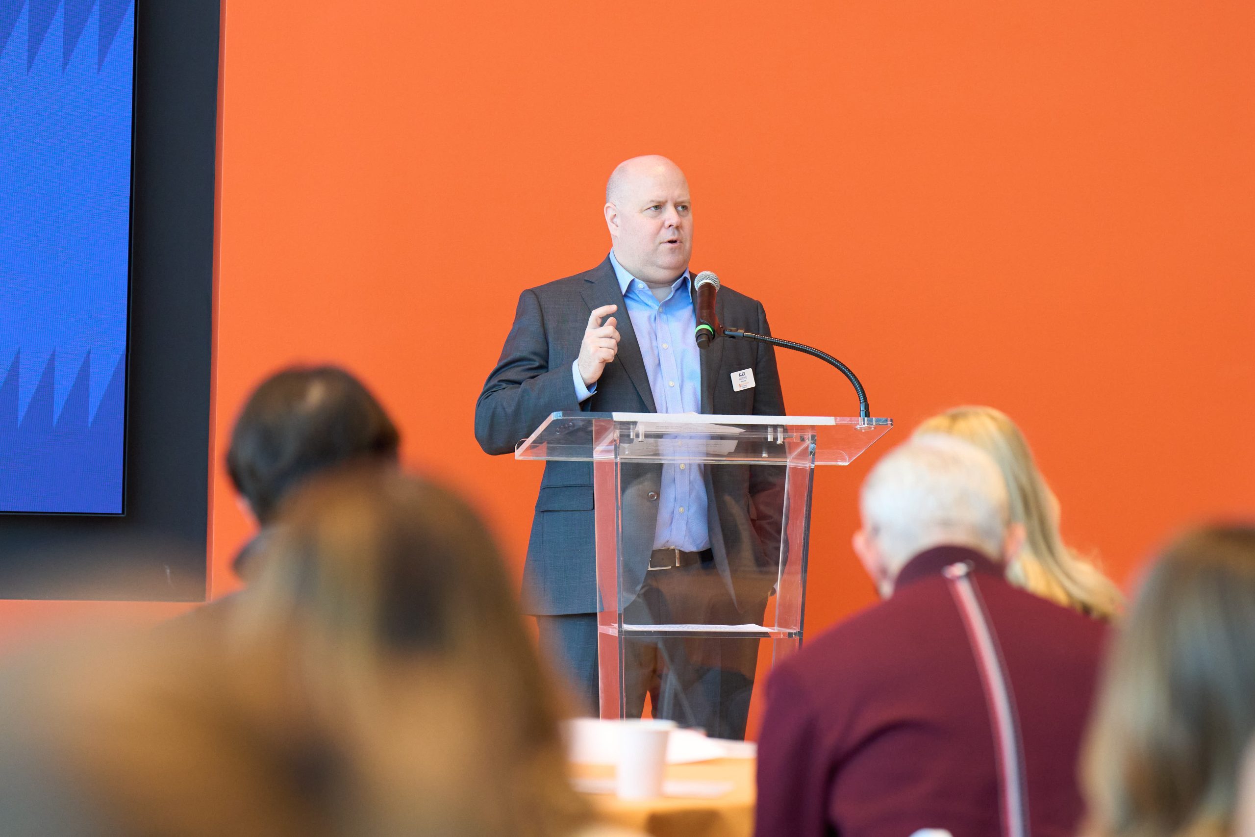 A person speaking at a podium in front of an audience with an orange background.