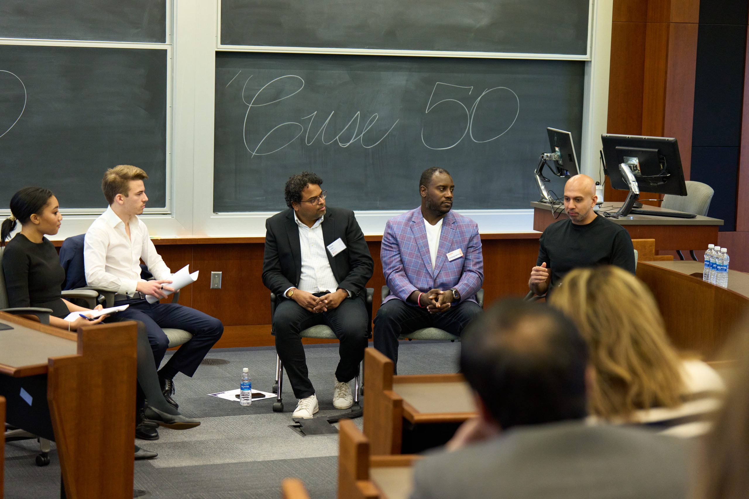 Five panelists are seated in front of a chalkboard with "Case 50" written on it, engaging with an audience in a university classroom.