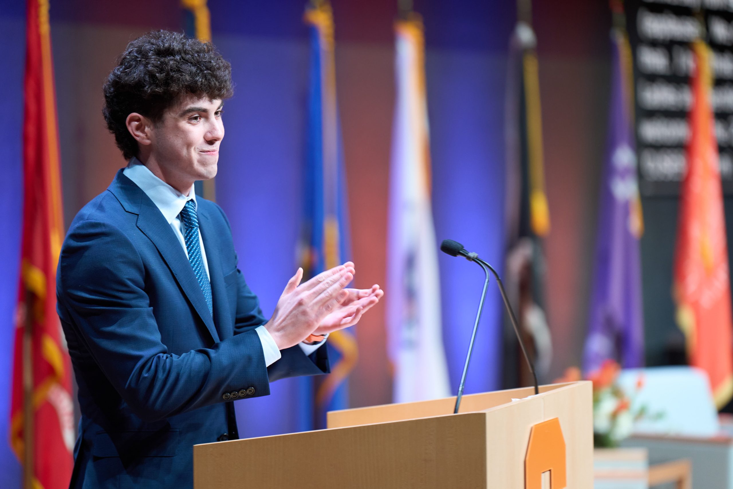 Person speaking at a podium with various flags in the background, at a formal event.