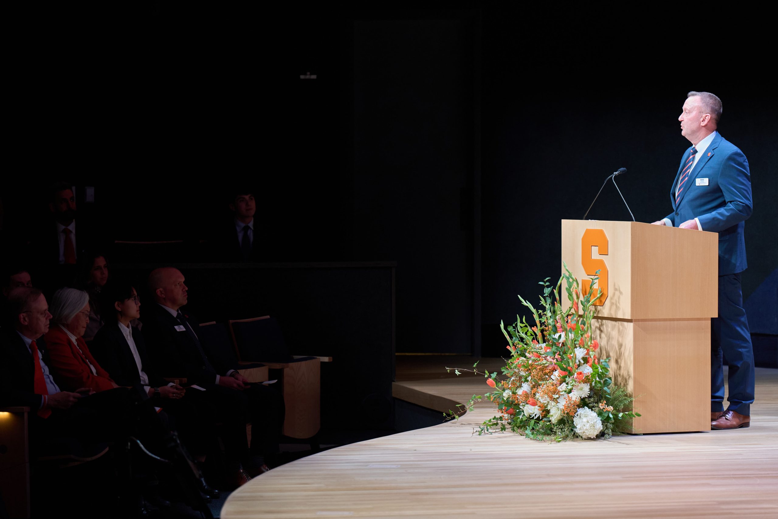 A person speaking at a podium with the Syracuse University logo, addressing an audience in a dimly lit auditorium.