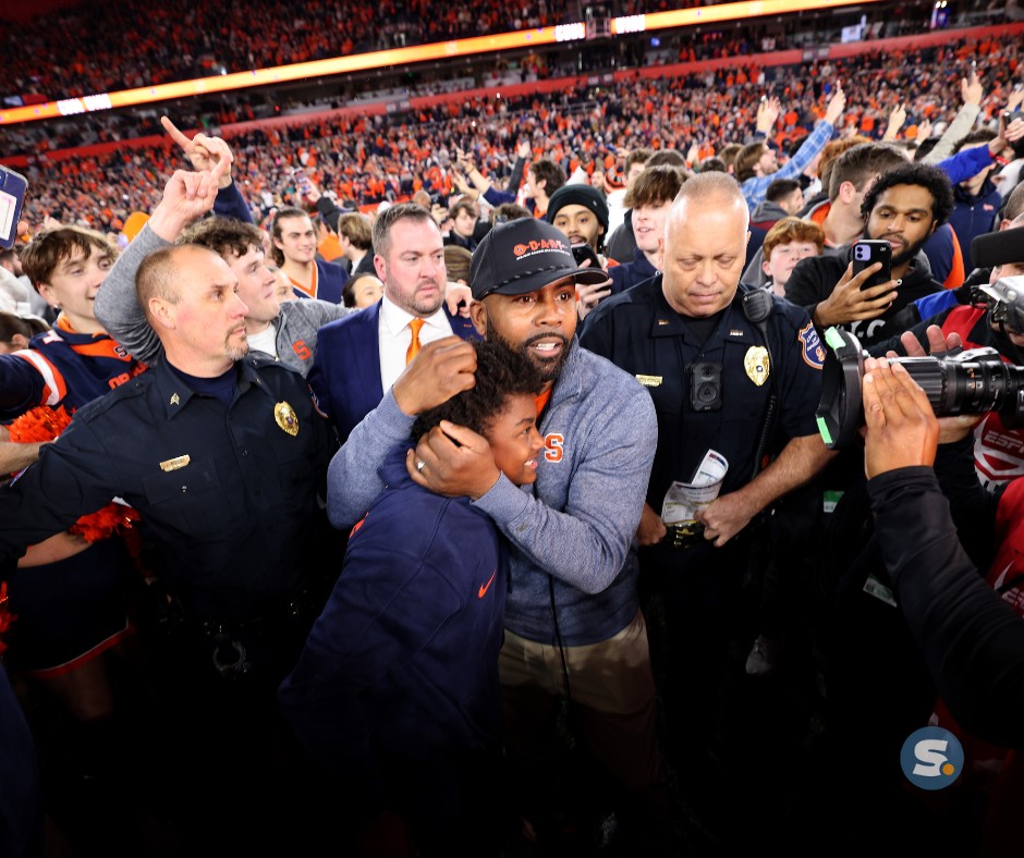 Head coach Fran Brown embracing his son in celebration surrounded by police and media during a crowded stadium event.