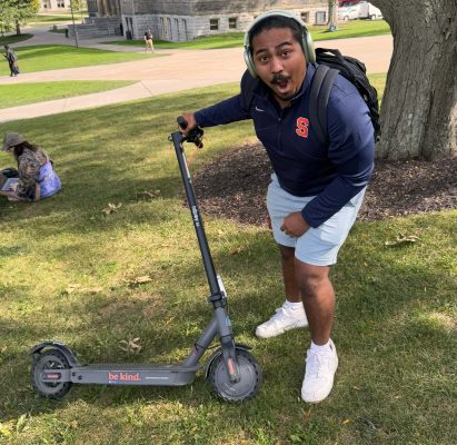 A person with a backpack stands excitedly beside a scooter labeled "be kind." in a park-like setting with trees and a stone building in the background. The individual is wearing a Syracuse University shirt.