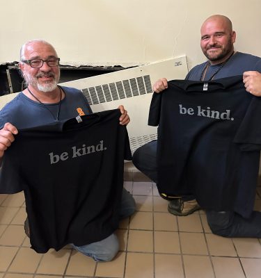 Two individuals smiling and holding up black T-shirts with the text "be kind" printed in white.