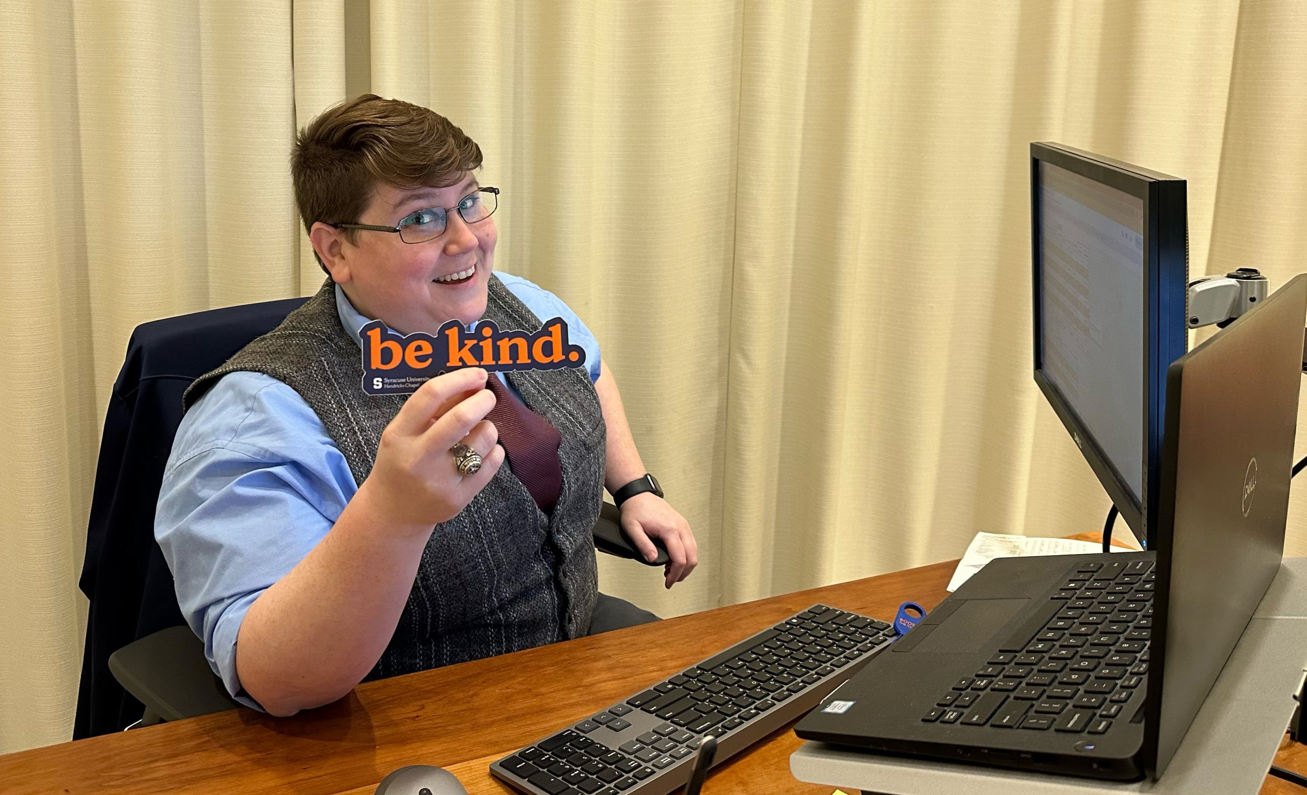 A person sitting at a desk in an office, holding a 'be kind' sign, and smiling while using a computer.