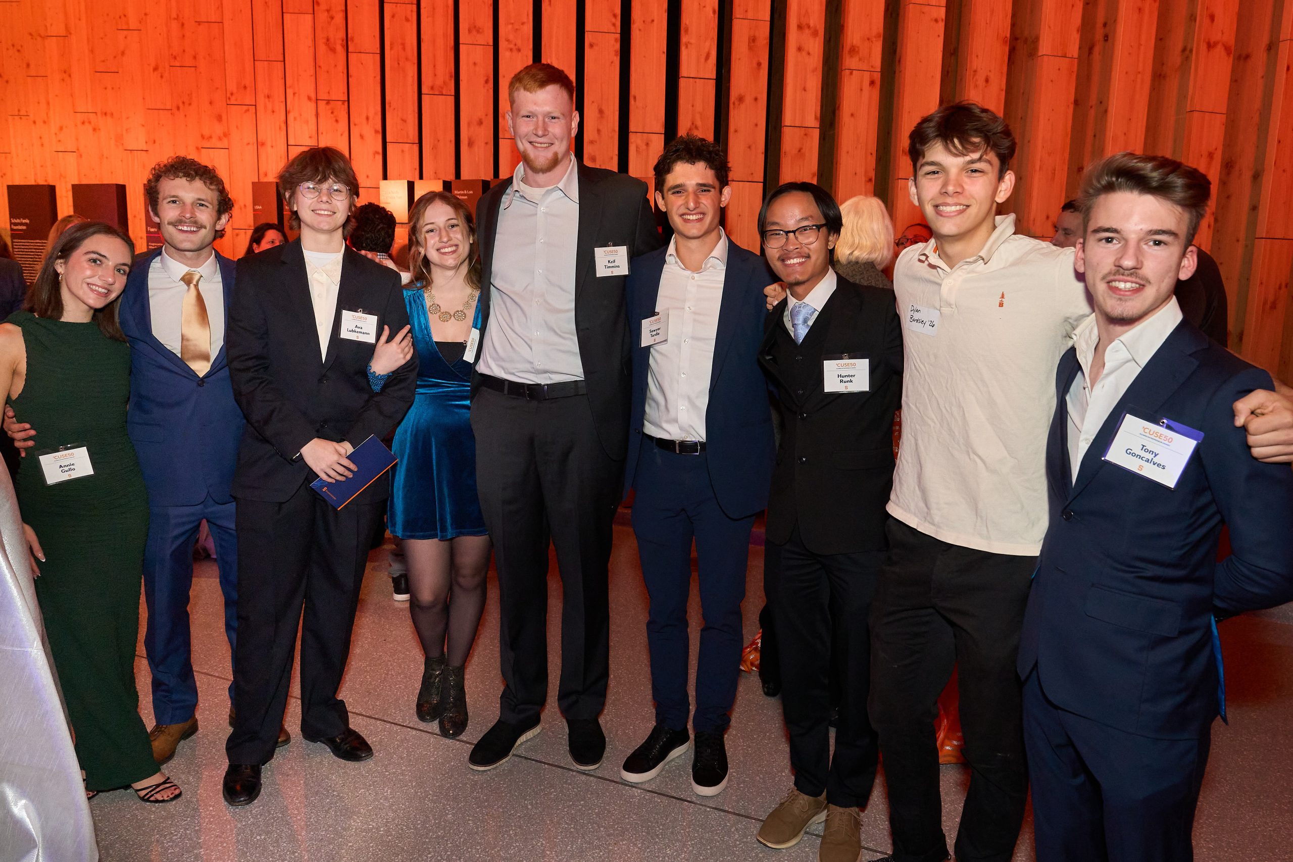 Group of eight individuals proudly posing at a formal event, smiling, dressed in business attire, with name tags.