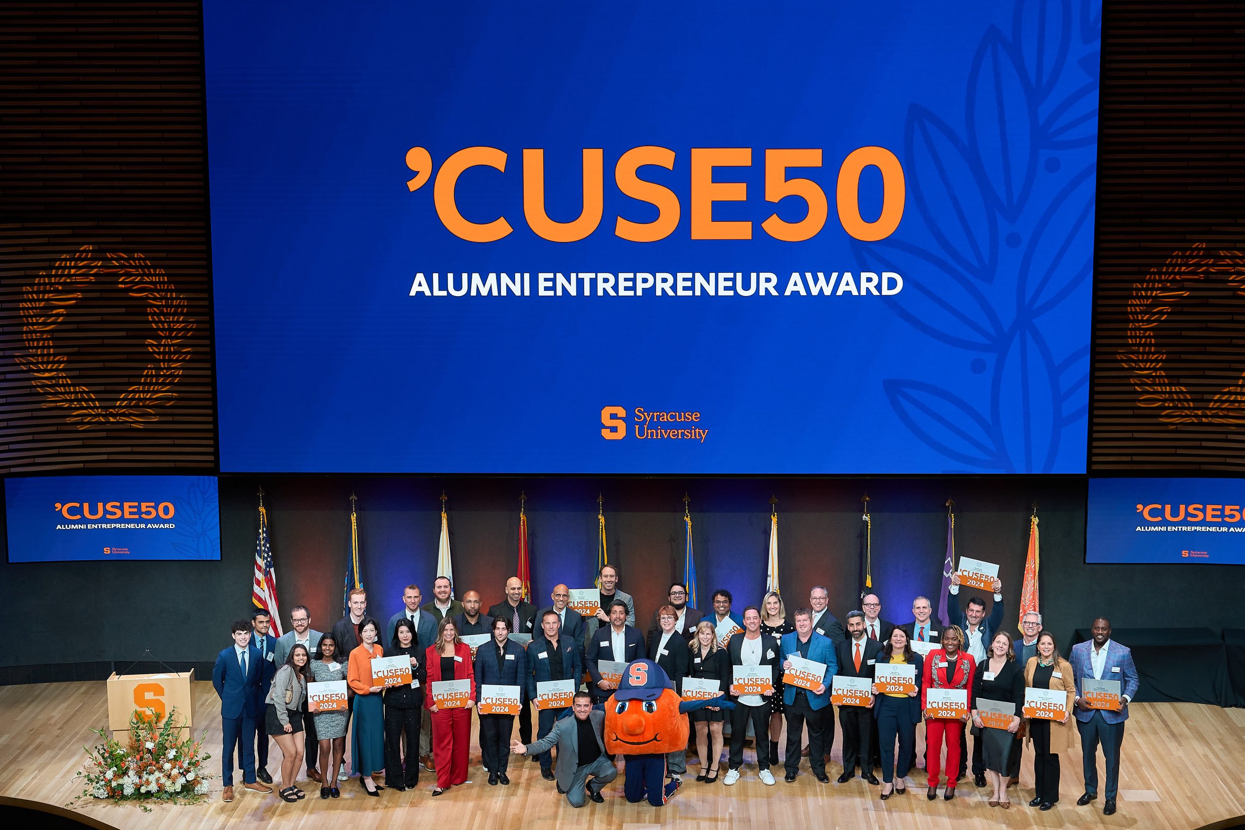 Group of individuals standing on stage holding flags, with a banner reading 'CUSE50 Alumni Entrepreneur Award' at Syracuse University event.