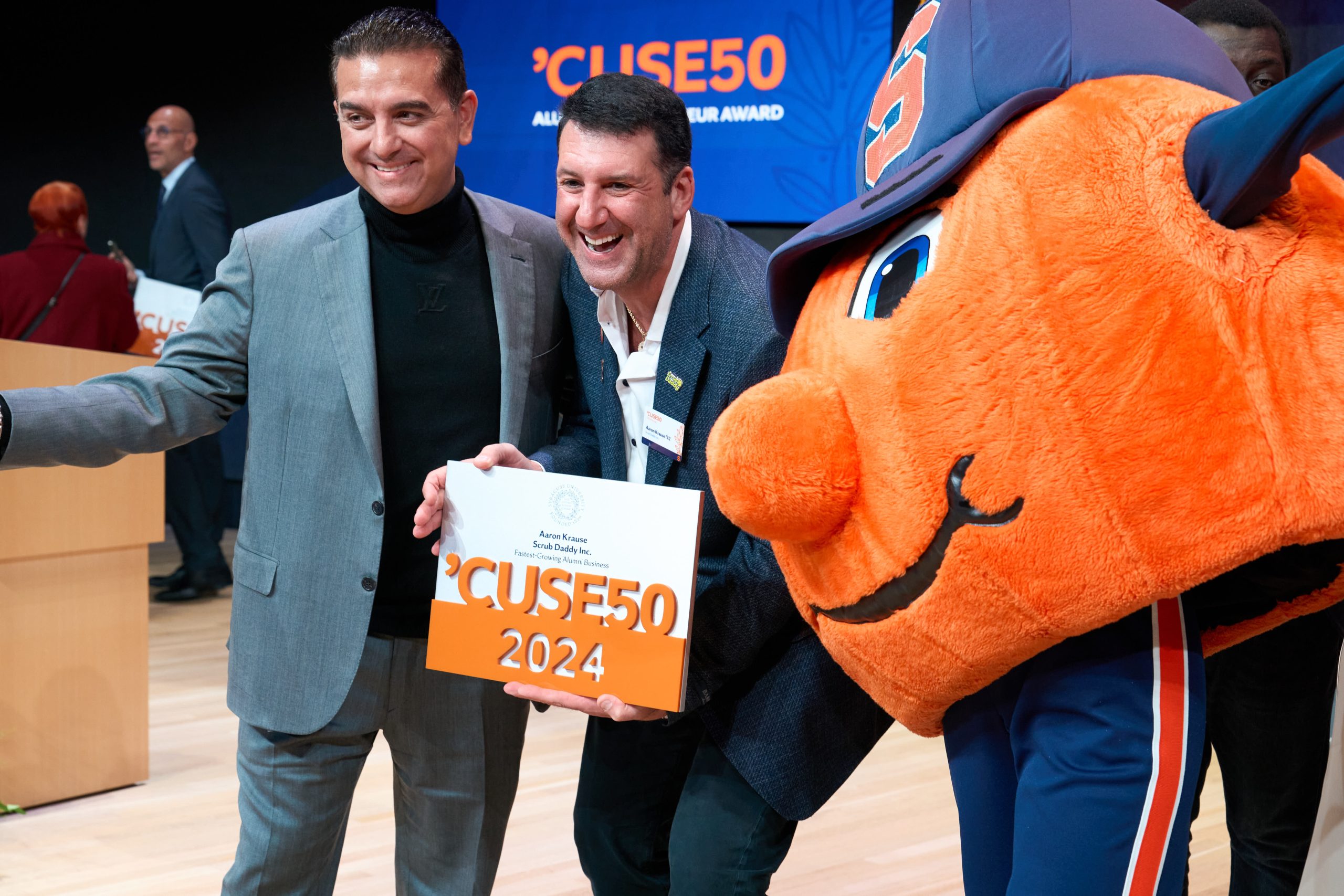 Two individuals holding an award certificate labeled "CUSE50 2024" stand smiling with Otto the Orange, the Syracuse University mascot, at an event.