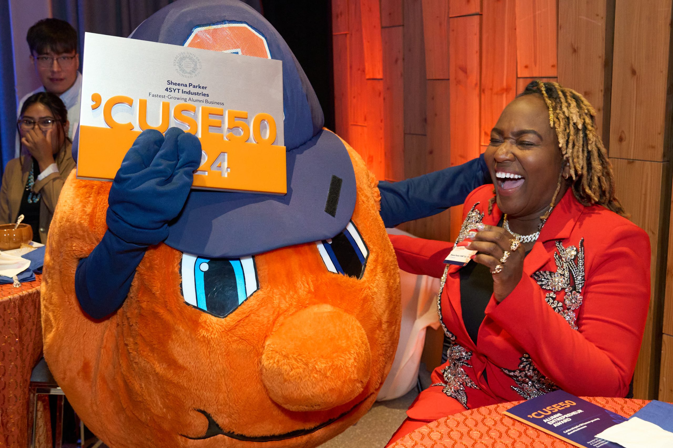 A person in business attire laughing joyfully next to a mascot resembling a large orange, wearing a hat and holding a Syracuse University 'Cuse50 sign, indoors at an event.