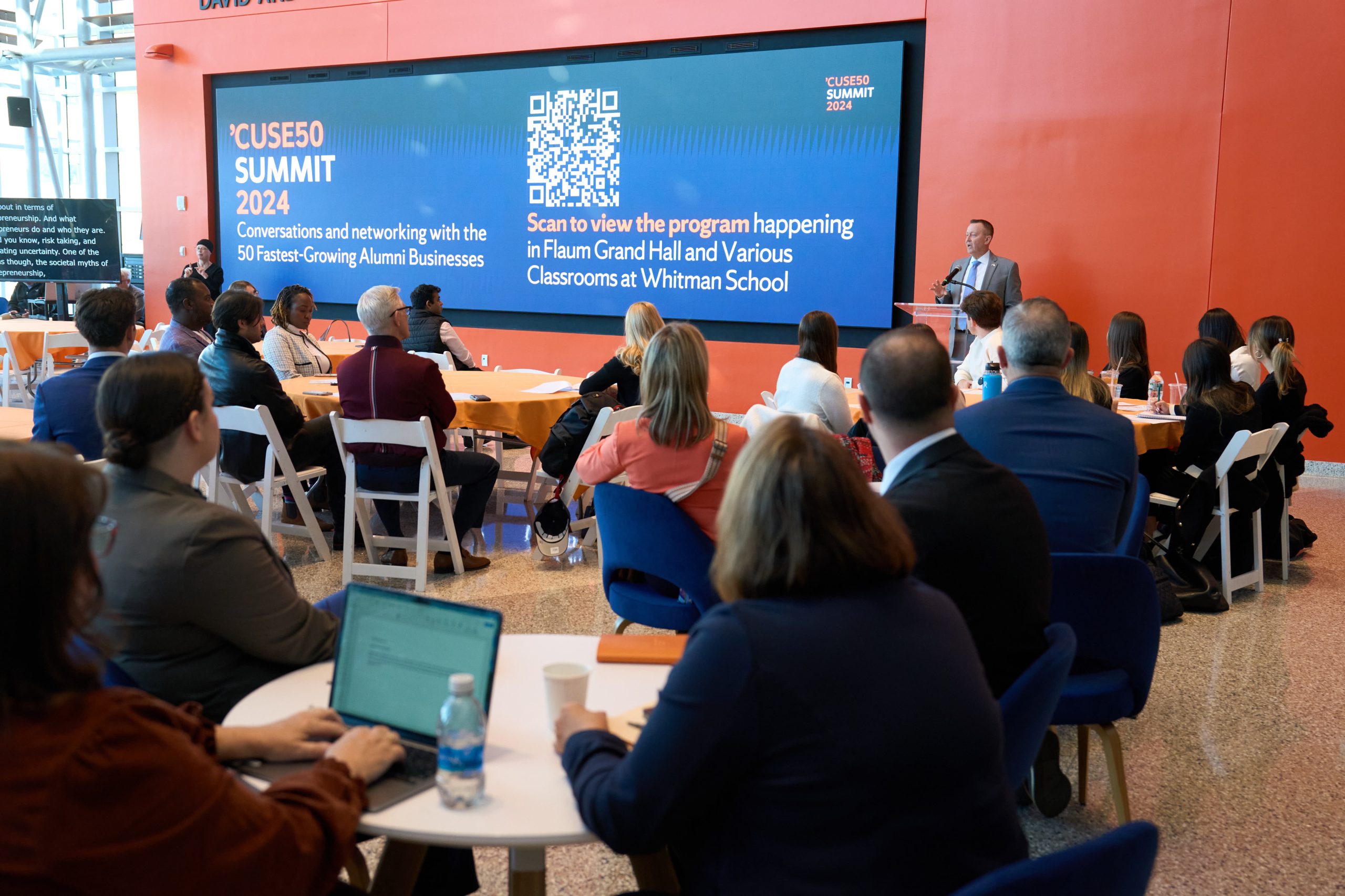 Attendees seated in a conference hall listening to a speaker at the CUSE50 Summit 2024, held in the Flavium Grand Hall at Whitman School. A digital screen displays a QR code for accessing the event program.