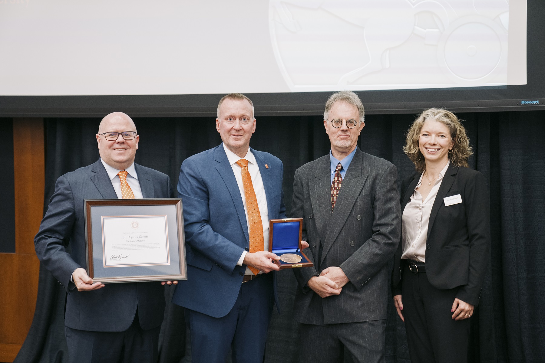 four people standing with one person holding a framed certificate and another person holding a box with a medallion