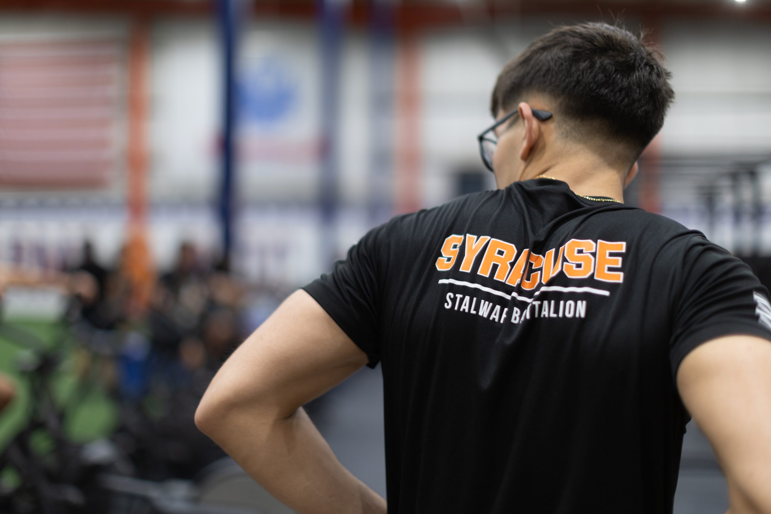 Person wearing a Syracuse Stalwart Battalion T-shirt, viewed from behind, in a gymnasium setting.