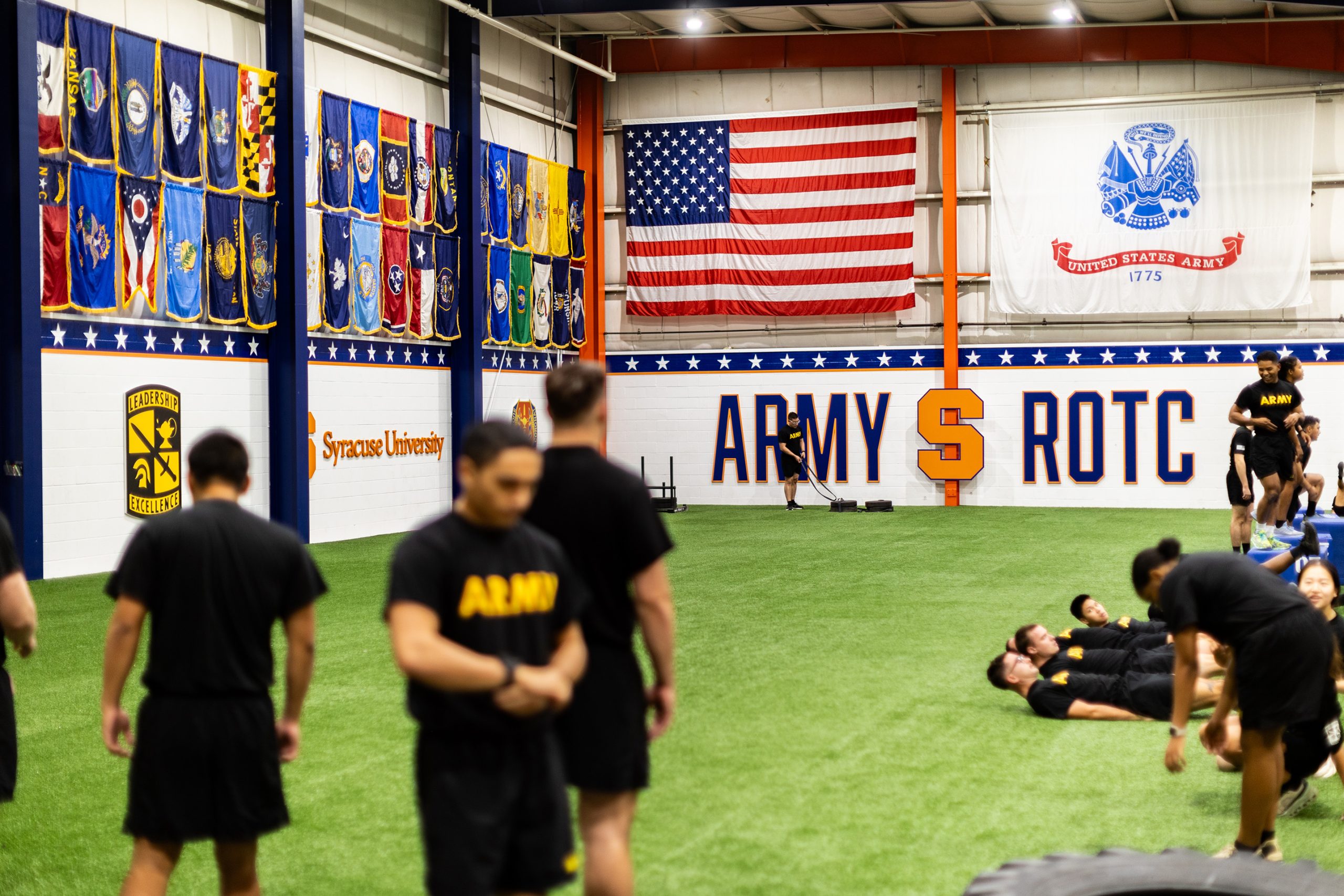 Individuals in physical training sessions at an indoor facility adorned with United States and Army ROTC flags, along with banners from Syracuse University.